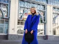 Uśmiechnięta blondynka w niebieskim płaszczu, trzymająca czarny kapelusz. Zimne kolory otoczenia podkreślają jej styl.