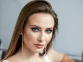 Brązowe włosy pięknie podkreślają niebieskie oczy. Modelka ma delikatny makijaż i długie, błyszczące kolczyki.