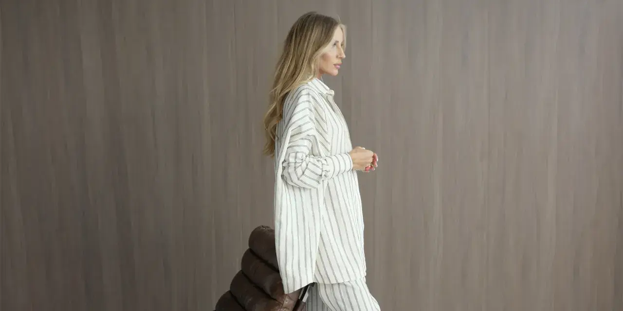 Elegancka kobieta w pasiastym, monochromatycznym stroju stoi obok brązowego fotela.