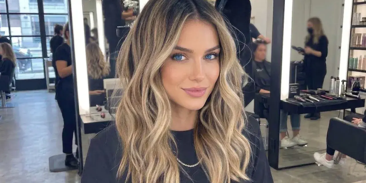 Kobieta z pięknymi, falowanymi włosami, blond z odrostem, ma hipnotyzujące niebieskie oczy. W tle salon fryzjerski.