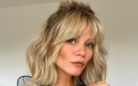Kobieta z niebieskimi oczami i piegami, z blond włosami i grzywką cieniowaną, ułożoną w luźny kok.