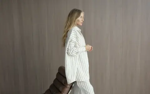 Elegancka kobieta w pasiastym, monochromatycznym stroju stoi obok brązowego fotela.
