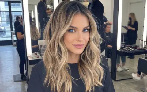 Kobieta z pięknymi, falowanymi włosami, blond z odrostem, ma hipnotyzujące niebieskie oczy. W tle salon fryzjerski.