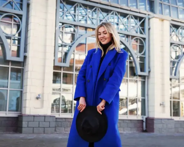 Uśmiechnięta blondynka w niebieskim płaszczu, trzymająca czarny kapelusz. Zimne kolory otoczenia podkreślają jej styl.