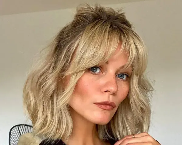 Kobieta z niebieskimi oczami i piegami, z blond włosami i grzywką cieniowaną, ułożoną w luźny kok.
