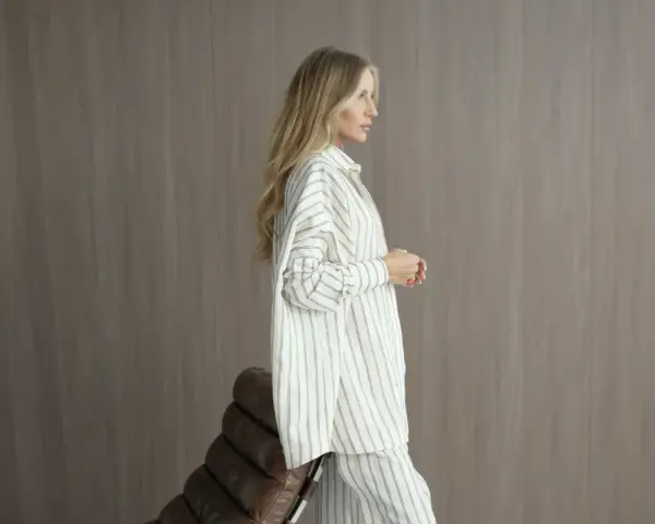 Elegancka kobieta w pasiastym, monochromatycznym stroju stoi obok brązowego fotela.