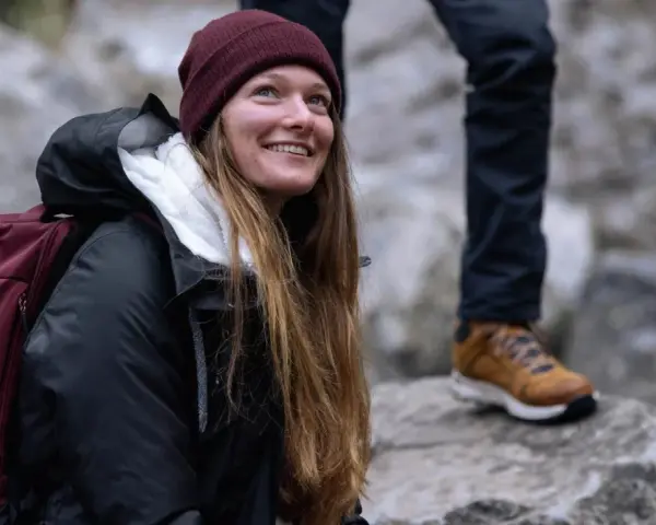 Uśmiechnięta kobieta w bordowej czapce i czarnej kurtce z bordowym plecakiem.