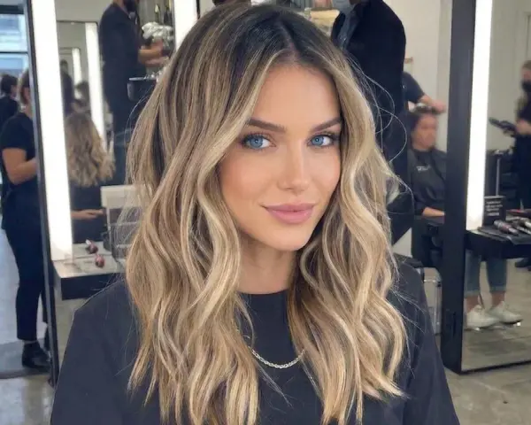 Kobieta z pięknymi, falowanymi włosami, blond z odrostem, ma hipnotyzujące niebieskie oczy. W tle salon fryzjerski.