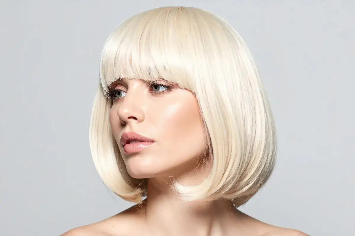 Blond fryzura paź bob z grzywką, podkreślająca delikatne rysy twarzy i błyszczące usta.