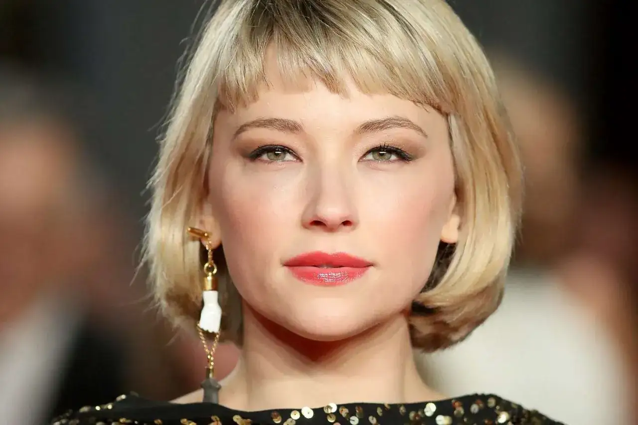 Kobieta z blond włosami i grzywką baby bangs, w eleganckiej sukni ozdobionej cekinami, z wyrazistym makijażem i oryginalną biżuterią.
