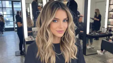 Kobieta z pięknymi, falowanymi włosami, blond z odrostem, ma hipnotyzujące niebieskie oczy. W tle salon fryzjerski.
